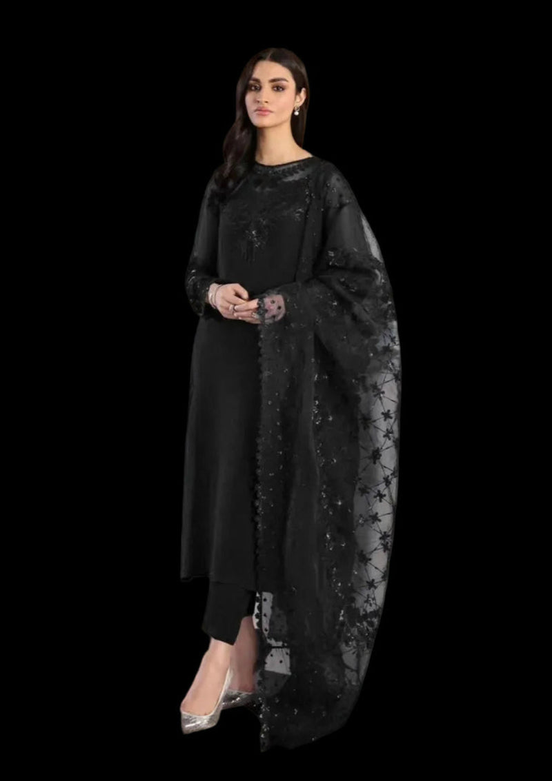 A woman models a black three-piece traditional outfit consisting of a long-sleeve kurta, matching trousers, and a sheer embroidered dupatta, set against an indoor wall with white flowers in the foreground.